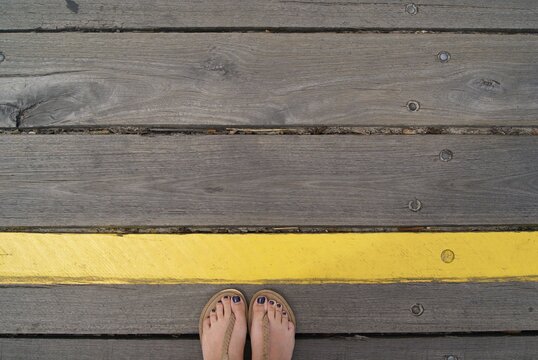 Overhead view of a woman in a pair of thong sandals standing behind a yellow line painted on wooden decking