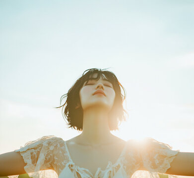 Young woman with eyes closed and arms outstretched in bright sunlight under a soft sky, freedom