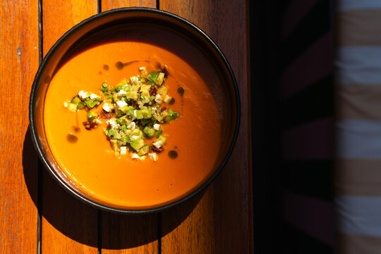 Overhead view of a bowl of Spanish salmorejo soup with avocado, feta cheese, quinoa and sun-dried tomato garnish on a wooden table in sunlight