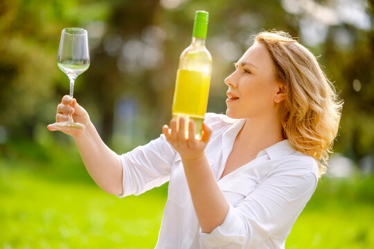 Blonde woman looking at white wine bottle and holding empty clear wineglass in park. Side view of elegant adult woman inspecting commercial beverage, sommelier concept