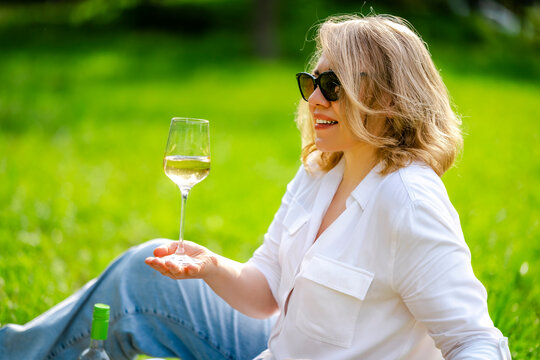 Smiling blonde woman holding Gewurztraminer wine glass at summer picnic park. Happy weekend recreation and social gathering concept.