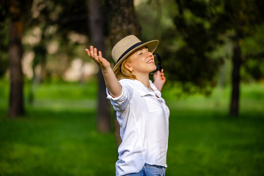 Happy adult woman in white shirt straw hat standing with open arms in sunny park. Freedom tranquility happiness wellness concept.