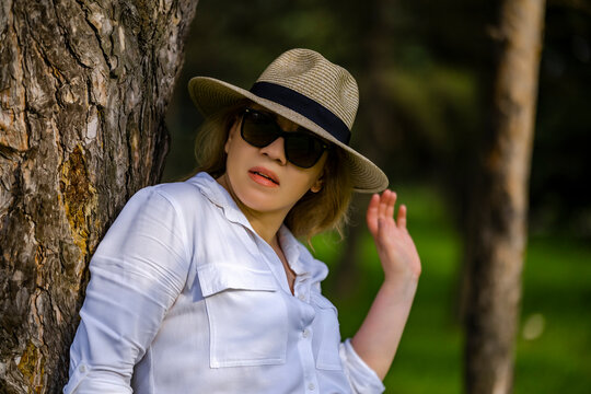 Beautiful adult woman in straw hat, sunglasses and white shirt leaning against tree trunk. Summer walk nature relaxation concept.