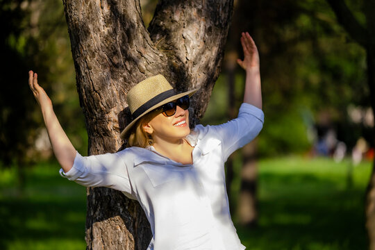 Happy woman in straw hat raising hands near tree in summer park. Freedom and mindfulness commercial concept.