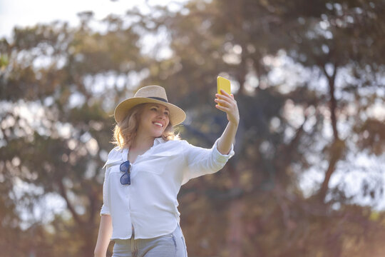 Smiling woman in straw hat taking selfie with yellow smartphone outdoor. Mobile communication and digital lifestyle commercial concept.