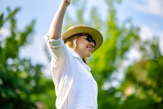 Cheerful woman in sunglasses raising hand in sunny summer park. Joyful emotion and energetic lifestyle commercial concept.