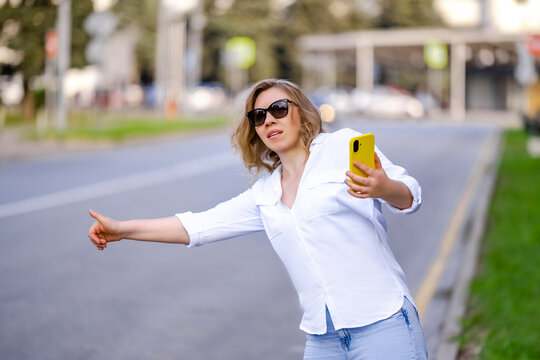 Anxious woman in sunglasses hitchhiking on roadside with yellow smartphone. Urban travel and transport breakdown commercial concept.