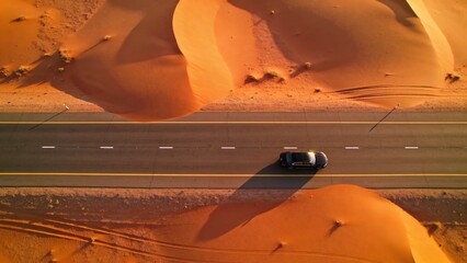 Naklejka premium Aerial View of a Black Car on a Desert Road Surrounded by Golden Sand Dunes
