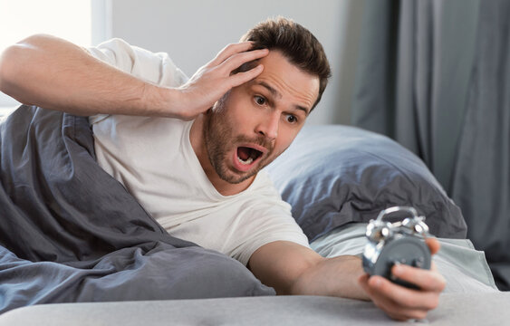 Overslept Man Holding Alarm Clock Waking Up Too Late For Work Lying In Bed At Home. Oversleeping Concept. Selective Focus