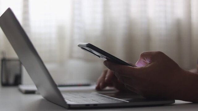 Close-up of man hand watching mobile phone while working on laptop at home office. Male finger typing and tapping on smartphone. Businessman using mobile apps, chatting, texting and scrolling.