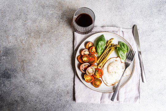 Overhead view of a plate of fermented asparagus, cherry tomatoes and a fried egg with a glass of fruit juice on a concrete table