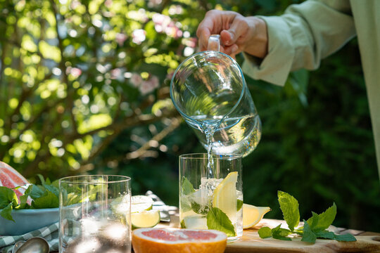 Close-up of a woman standing by a garden table pouring water into a glass with fresh lemon and mint leaves