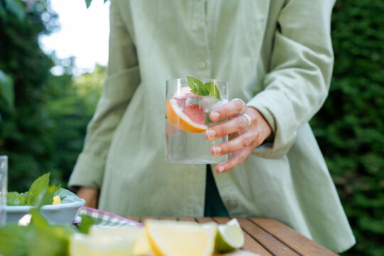 Close-up of a woman standing in front of a garden table holding a refreshing glass of water with fresh grapefruit and mint leaves