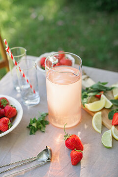 Close-up of a pitcher of strawberry lemonade, two glasses, fresh strawberries, limes and mint leaves on a garden table on a summer day
