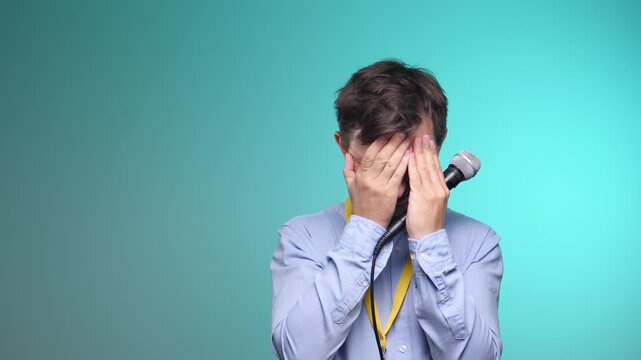 Young man covering face with hands and holding a microphone, showing visible signs of nervousness and anxiety before or during a public speaking event against a blue background