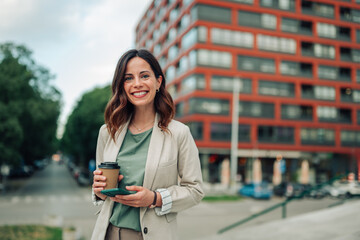 Businesswoman standing outdoors holding smartphone and coffee, smiling confidently in urban setting...