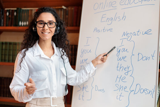 Positive young school teacher working from home, teaching English language grammar rules subject to students online. Woman pointing at whiteboard, talking with headphone.