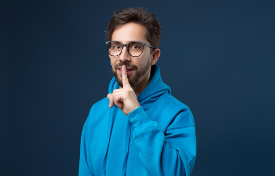 Make Silence. Portrait Of Young Man Showing Shh Gesture At Camera, Millennia Guy In Eyeglasses Holding Finger Near Lips, Gesturing Hush While Standing Isolated On Blue Studio Background