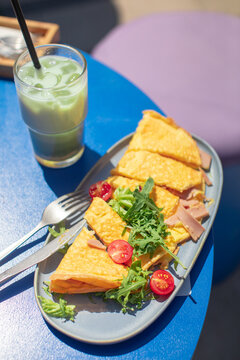 Close-up of a ham and cheese omelette with rocket and tomato garnish and an iced matcha latte on a table