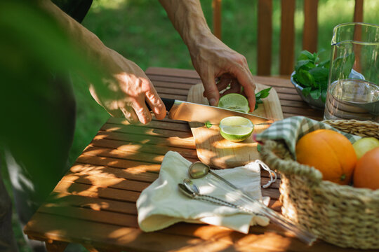 Close-up of a man standing by a garden table cutting a fresh lime to make a citrus lemonade drink on a summer day