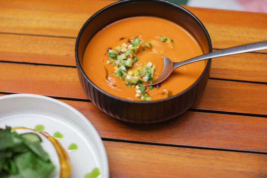 Close-up of a bowl of Spanish salmorejo soup with avocado, feta cheese, quinoa and sun-dried tomato garnish on a wooden table in sunlight next to an open-faced toasted egg and cheese bun