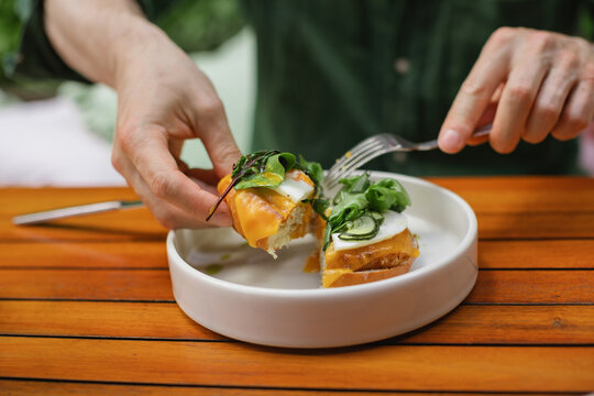 Close-up of a man sitting at a table eating a halved open-faced toasted egg and cheese bun with salad