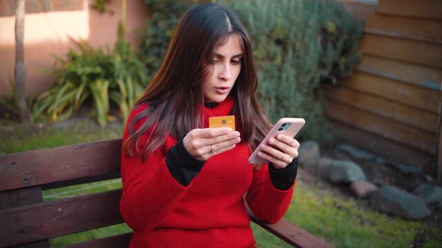 Young Hispanic woman in her home garden trying to make an online purchase with her credit card on a smartphone, becoming frustrated after a failed payment or transaction.
