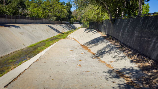 A concrete storm drain channel or urban river bed runs beneath green trees and a black fence under a bright clear blue sky with a trickle of water flowing over moss and algae