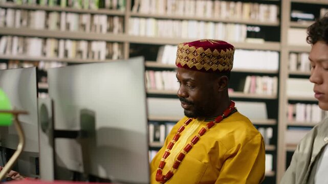 Adult student in traditional clothing work at desktop along shared library table with books, keyboards and monitors, for digital learning and academic training.