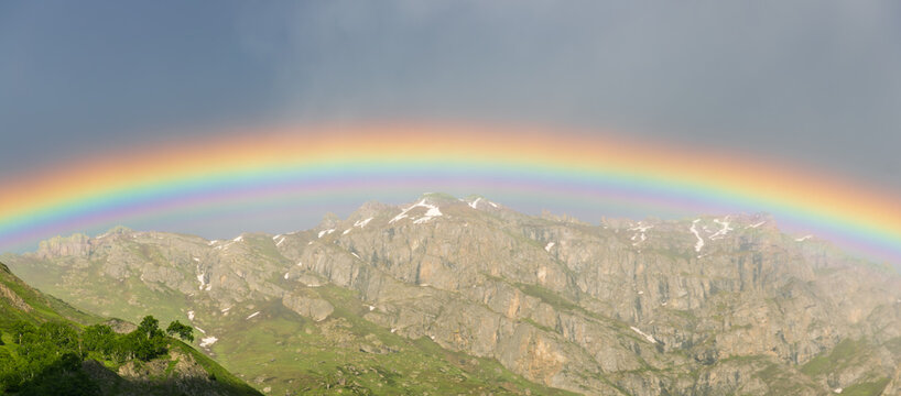 A bright horizontal rainbow perfectly divides the dark storm clouds from rugged rock peaks in ultra wide ground landscape, isometric framing of a rainbow splitting a stormy sky and Rocky Mountains