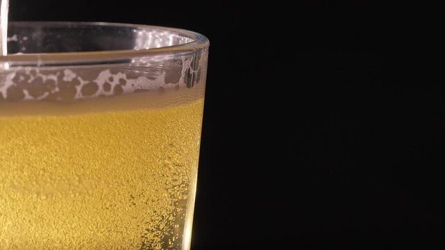 Golden beer pouring into clear glass with rising foam and sparkling bubbles creating refreshing lager drink on black background.