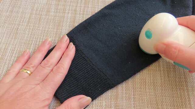 Closeup of female hands carefully using an electric lint remover to clean pills and fuzz from a blue sweater. Housework and garment care concept.