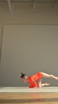 A little Caucasian girl gymnast in an orange leotard demonstrates her skills on a white background. 