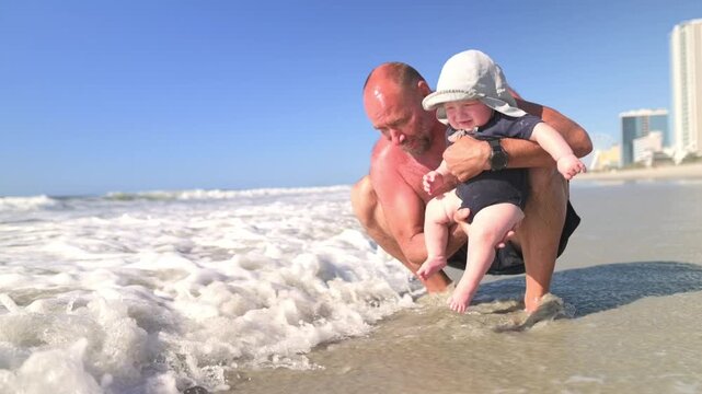 Man with shaved head crouches in shallow ocean water, holding a smiling baby in a sun hat, as waves splash around them and a beachside cityscape is visible in the background