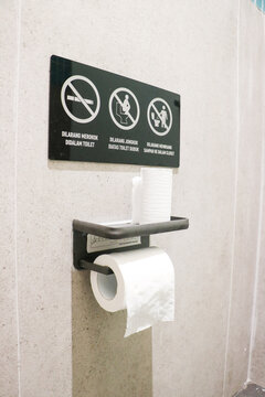 A black public restroom sign with explicit rules mounted above a metal toilet paper holder