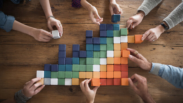 Diverse hands collaboratively building a colorful block graph on a wooden table, top view, ideal for business growth, teamwork and data visualization concepts