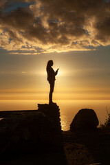 Fototapeta premium A silhouette of a woman stands on a cliff edge, looking at her phone, as the sun sets over the ocean in Big Sur, California.