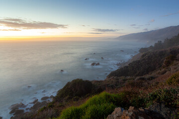 Obraz premium The Pacific Ocean meets the rugged coastline of Big Sur, California, at dusk. The soft light of sunset casts a serene glow over the misty waters and rocky shores.