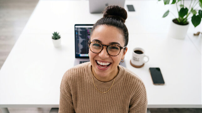High angle shot of a happy young woman with glasses and a bun hairstyle smiling at the camera while working at her desk in an office