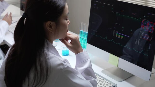 Focused Asian woman scientist typing on computer keyboard, analyzing clinical laboratory data, pointing at screen