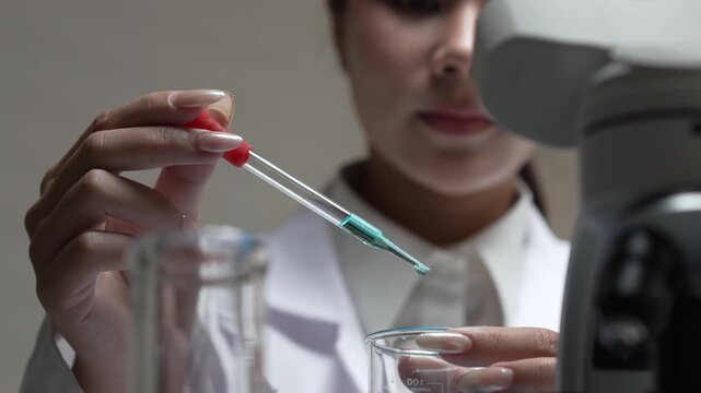 Focused female scientist conducts biotech research, using pipette with liquid, beaker and microscope in laboratory