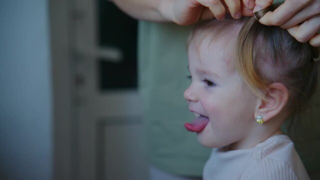 Young girl sticks tongue out while adult styles her hair. She wears pearl earring and light sweater. Scene shows playful moment during hair styling. Footage suitable for family, beauty