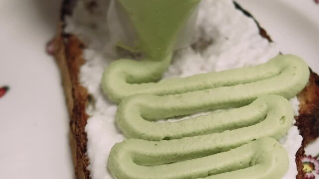 Cooking bruschetta. Tasty appetizer. Chef hand squeezing pastry bag for piping green cream on plated toasted bread with soft white spread.