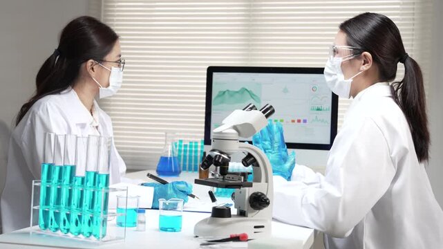 Focused asian female scientist and researcher analyzing clinical medical sample with computer, microscope inside laboratory