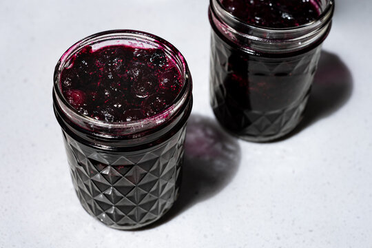 Glass jars of homemade blueberry jam, or dessert topping, sitting on white stone kitchen counter, delicious summer food treat
