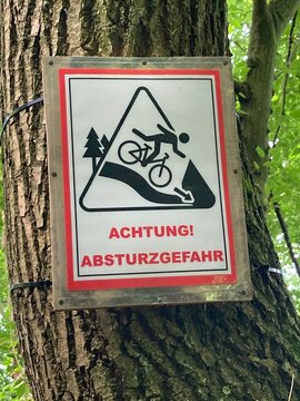 Warnung - Schild Absturzgefahr f&uuml;r Fahrradfahrer  an einem Baum im Wald am Abhang
