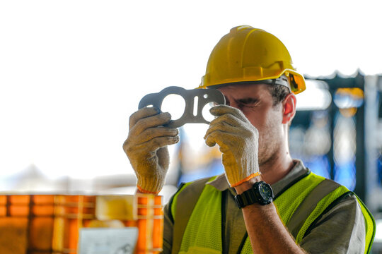 Young male warehouse technician in a hard hat and reflective safety vest holds a precision gasket up to check its openings in a vehicle parts factory with industrial machinery and steel rac.