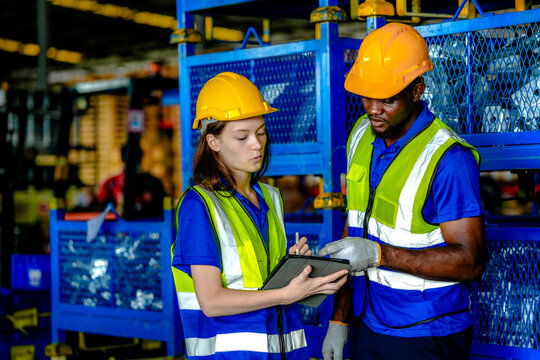 African male warehouse worker and young Caucasian female colleague in safety vests record contents of industrial mesh containers in a vehicle parts factory with industrial machinery and.