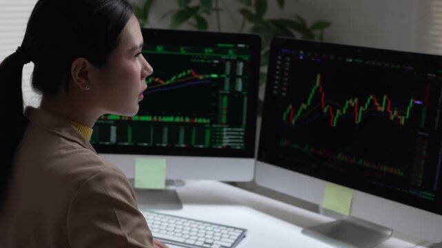 Focused asian woman trader analyzing complex stock market graph on computer monitor in dark office late at night