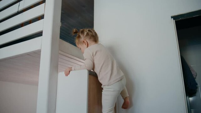 Toddler climbs a bunk bed ladder. Child leans forward, gripping the rail. Barefoot and focused, she pushes upward. She reaches the top step, turning to climb further